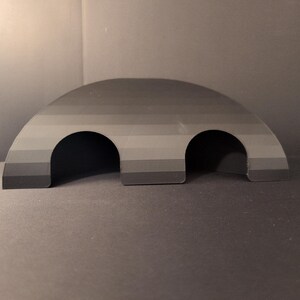 Puede incluir: Una estructura de escondite gris en forma de semicírculo que presenta un degradado de bandas horizontales que van del gris oscuro al gris claro. El objeto tiene dos aberturas arqueadas en la base, que dan acceso al espacio interior, con un acabado liso.