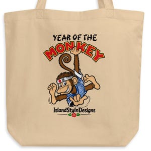 May include: A beige tote bag featuring a graphic of a brown monkey wearing a blue patterned shirt and a white headband with a red circle. The text reads YEAR OF THE MONKEY and IslandStylnDesigns, with a small red hibiscus flower icon below.