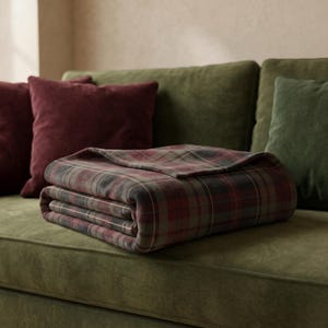 May include: A folded throw blanket featuring a plaid pattern in shades of dark red, olive green, and charcoal gray with thin white intersecting lines. The fabric has a soft, matte texture and is neatly layered on a green upholstered sofa.