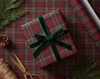 May include: A rectangular gift box wrapped in dark red, green, and gold plaid paper, tied with a dark green velvet ribbon. The scene includes a roll of matching wrapping paper, gold scissors, a spool of twine, and evergreen sprigs with pinecones.
