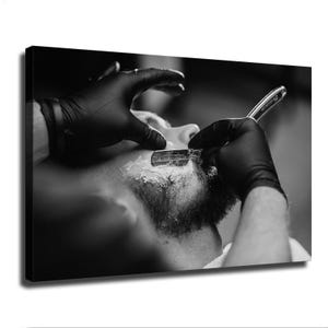 Può includere: Una stampa su tela in bianco e nero che raffigura un primo piano di una rasatura con rasoio a mano libera. Un paio di mani con guanti neri tiene un rasoio metallico contro una mascella barbuta coperta di schiuma da barba bianca, catturando il movimento.