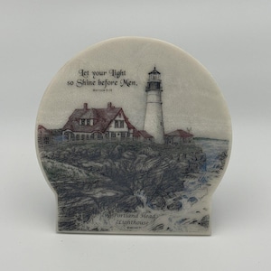 May include: A semi-circular stone plaque featuring a detailed illustration of the Portland Head Lighthouse on a rocky coastline. The text reads: Let your light so Shine before Men. Matthew 5:16 and Portland Head Lighthouse.