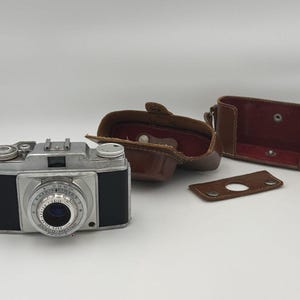 May include: An Ansco Memar camera with a silver and black body, featuring a Pronto lens. The camera is accompanied by a brown leather case that is separated into three distinct pieces, including the main body cover and a small lens cap.