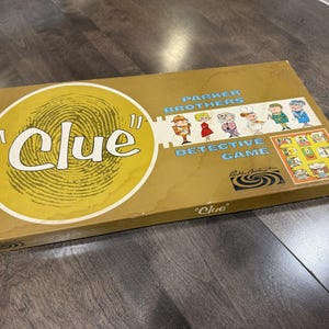 May include: A rectangular Clue board game box with a gold-toned cover featuring a large white fingerprint graphic, the text Clue, Parker Brothers Detective Game, and illustrations of six characters. A small game board image is also printed on the box.