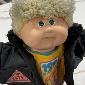 May include: A Cabbage Patch Kids doll with blonde yarn hair and green eyes, wearing a black jacket with a triangular logo patch, over a yellow shirt featuring the text 199 SOUTH U.S.A. and a red and green checkered pattern on the front.