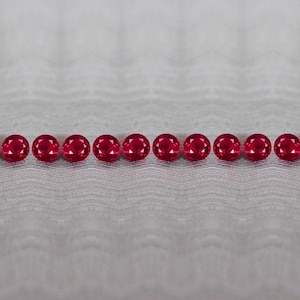 Puede incluir: Una fila horizontal de diez piedras preciosas redondas, facetadas y de color rojo intenso. Cada piedra presenta un corte brillante con múltiples facetas triangulares que reflejan la luz, creando un aspecto brillante en los rubíes.