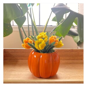 May include: A bright orange ceramic pumpkin-shaped planter containing a bouquet of fuzzy yellow and orange pipe cleaner flowers with green stems. The planter has a smooth, glossy finish and vertical ridges, creating a rounded, segmented appearance.