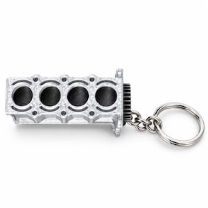 May include: A silver-colored metal keychain featuring a miniature four-cylinder engine block replica. The engine block has four circular openings with dark interiors and a small gear attached to one end, connected to a standard circular metal key ring.