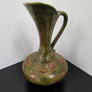 May include: A ceramic pitcher featuring a mottled green and reddish-brown glaze. The vessel has a wide, rounded base, a slender neck, and a curved handle. A decorative, raised petal-like pattern encircles the base of the neck, and the rim is flared.