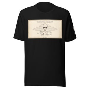 Op de afbeelding: Een zwart t-shirt met korte mouwen en een rechthoekige grafische print met de titel THE XENO ARCHIVE - SPECIMEN CARD. De print toont een grijze alien voor een vliegende schotel met labels die naar kenmerken wijzen, waaronder Ovis Cinereus - Grey - Origin Unknown.