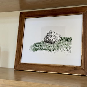 May include: A framed art print featuring a black and white ink illustration of a large, textured rock resting in a patch of green grass. The artwork is set against a light gray square background and enclosed in a dark brown wooden frame with gold trim.