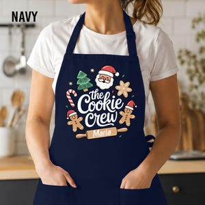 May include: A navy blue apron featuring a white graphic print that says "the Cookie Crew" with a rolling pin labeled "Maria" underneath. The design includes a Santa Claus face, two gingerbread people, a Christmas tree, and a candy cane.