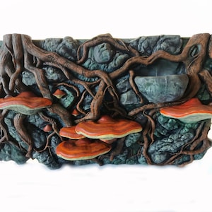 Puede incluir: Una escultura de pared rectangular que presenta una superficie rocosa de color verde oscuro texturizada con raíces de árbol marrones extendidas. Varios hongos de estante naranjas y rojos están adheridos, junto a una pequeña cavidad en forma de cuenco.