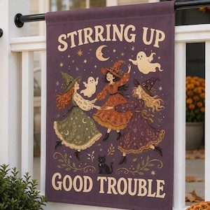 May include: A rectangular purple garden flag featuring the text "STIRRING UP GOOD TROUBLE" in cream lettering. The illustration depicts three figures in witch hats and dresses dancing in a circle with two small ghosts and a black cat.