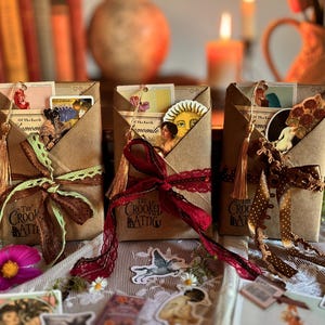 May include: Three rectangular brown paper packages labeled The Crooked Attic, each tied with a decorative ribbon and lace. The packages contain assorted paper ephemera, including vintage-style illustrations, sun motifs, and floral cutouts.