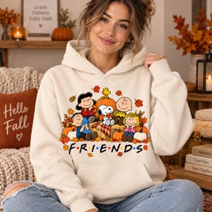 Könnte beinhalten: Ein cremefarbener Kapuzenpullover mit einem Grafikdruck der Peanuts-Figuren, die mit Kürbissen und Herbstblättern sitzen. Der Text FRIENDS ist unter den Figuren in einer bunten Schriftart gedruckt, umgeben von verstreutem Herbstlaub.