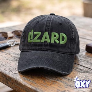 Może przedstawiać: Czarna czapka z daszkiem o postarzanym wykończeniu z postrzępionymi krawędziami daszka. Słowo LIZARD jest wyhaftowane limonkowymi wielkimi literami na przednim panelu. Czapka posiada guzik na górze oraz przeszywane oczka zapewniające wentylację.