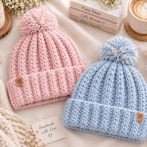 May include: Two knit beanies, one light pink and one light blue, each featuring a ribbed texture, a matching pom-pom on top, and a small brown rectangular tag on the folded brim with a black heart icon printed on the center of the tag.
