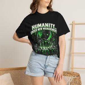 Könnte beinhalten: Ein schwarzes T-Shirt mit einem Grafikdruck einer Roboterfigur, die eine Menschenmenge mit grünen Laserfäden kontrolliert. Der Text "HUMANITY... UNDER NEW MANAGEMENT!" steht in weißen und grünen Buchstaben vor einem dunklen, futuristischen Hintergrund.