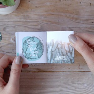 May include: A small, open book with a blue and green illustration of the world on the left page and a photo of a rocky landscape on the right page. The text on the left page reads "Go where you feel most alive". The text on the right page reads "The Burren County Clare".