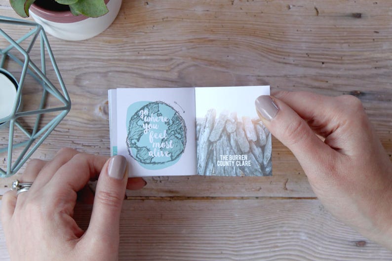 May include: A small, open book with a blue and green illustration of the world on the left page and a photo of a rocky landscape on the right page. The text on the left page reads "Go where you feel most alive". The text on the right page reads "The Burren County Clare".