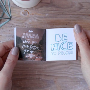 May include: A small, open book with a motivational quote on each page. The left page features a landscape scene with the quote "Do the best you can until you know better, when you know better, you do better." The right page has the quote "Be nice to people" in blue text.