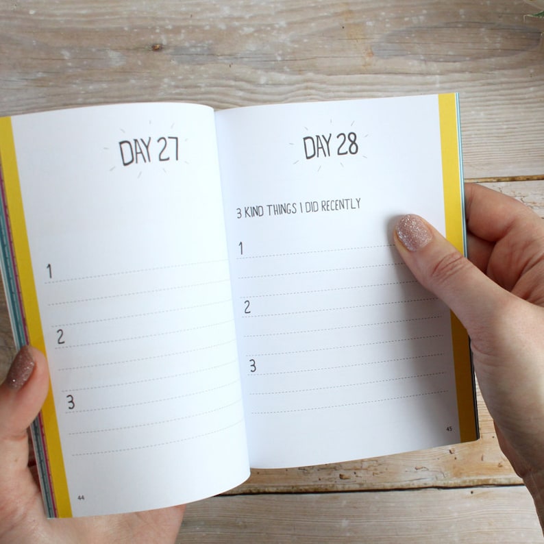 May include: An open journal with a yellow and white striped cover. The pages are numbered 44 and 45. The text on the page reads "Day 27" and "Day 28". The right page has the heading "3 Kind Things I Did Recently" with three lines for writing.