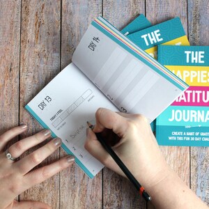 May include: A turquoise blue and yellow gratitude journal with the text "The Happiest Gratitude Journal" and "Create a habit of gratitude with this fun 30 day challenge". A hand is writing in a smaller turquoise blue journal with the text "Day 13" and "Day 14".