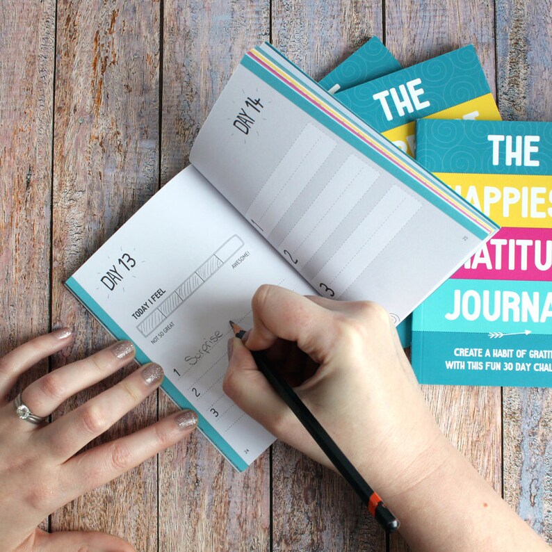 May include: A turquoise blue and yellow gratitude journal with the text "The Happiest Gratitude Journal" and "Create a habit of gratitude with this fun 30 day challenge". A hand is writing in a smaller turquoise blue journal with the text "Day 13" and "Day 14".
