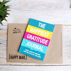 May include: A brown paper envelope with a "Happy Mail" stamp and a small, colourful journal titled "The Happiest Gratitude Journal: Create a Habit of Gratitude with this Fun 30 Day Challenge".