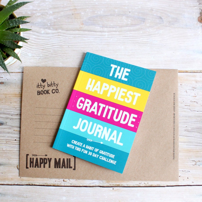 May include: A brown paper envelope with a "Happy Mail" stamp and a small, colourful journal titled "The Happiest Gratitude Journal: Create a Habit of Gratitude with this Fun 30 Day Challenge".