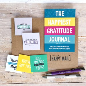 May include: A brown paper envelope with a colorful gratitude journal, four small motivational cards, and two pens. The journal has the text "The Happiest Gratitude Journal" and "Create a habit of gratitude with this fun 30 day challenge". The cards have positive messages like "You've got this" and "You are capable of amazing things". The pens have the text "Be kind to yourself" and "You're doing great".