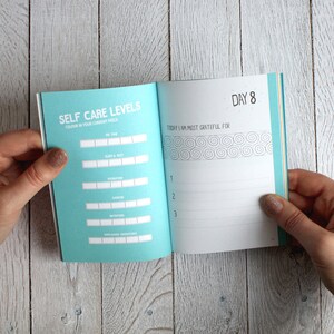 May include: An open journal with a blue cover and white pages. The left page has the title "Self Care Levels" and a chart to colour in. The right page has the title "Day 8" and the text "Today I am most grateful for".