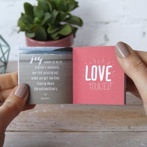 May include: A small pink and gray book with the text "joy comes to us in ordinary moments. we risk missing out when we get too busy chasing down the extraordinary." and "LOVE YOURSELF" on the front and back covers.