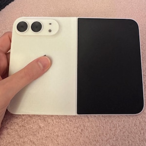 May include: A foldable smartphone featuring a two-tone design with a white left panel and a black right panel. The white section includes a horizontal camera module with two circular lenses and a small flash, while the device has a flat, rectangular shape.