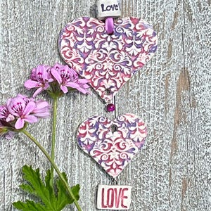 May include: Two pink and purple ceramic hearts with a white damask pattern, hanging from a pink ribbon with a "Love" charm. The hearts are hanging against a whitewashed wood background.