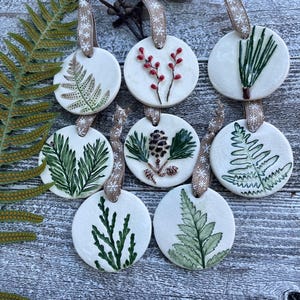 May include: A collection of round, white ceramic ornaments with botanical designs. Each ornament features a different hand-painted plant, including ferns and berries, with a burlap ribbon for hanging. The ornaments are arranged on a weathered wooden surface.