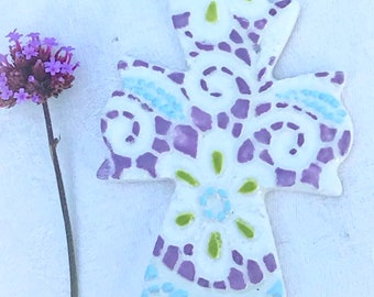 May include: A white ceramic cross with a purple, green, and blue mosaic design. The cross is hanging from a purple ribbon.