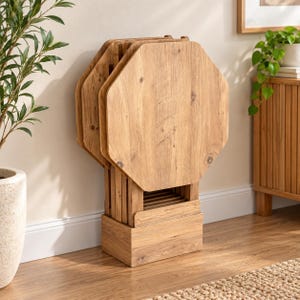 May include: A set of four light brown wooden snack tables with octagonal tops, neatly stored in a matching rectangular wooden stand. The wood features a visible natural grain pattern, and the entire unit is designed for compact vertical storage.