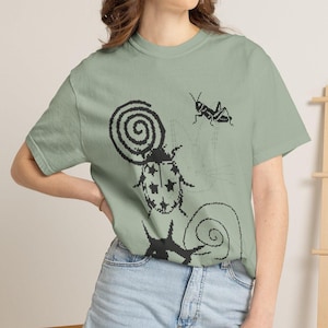 May include: A sage green short-sleeved t-shirt featuring a black pixelated graphic print. The design includes a large beetle with star-shaped markings, a spiral shape, a grasshopper, and a faint outline of a cat face in the background.