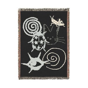 May include: A black woven throw blanket featuring a white graphic design of a ladybug, a grasshopper, a spiral, and an eye. The blanket has a fringed edge on the top and bottom, with a multi-colored thread detail visible along the side borders.