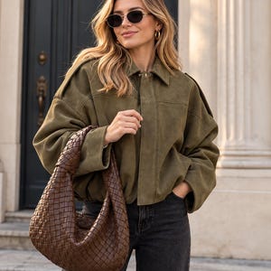 Puede incluir: Una chaqueta verde oliva con cuello y mangas largas, combinada con vaqueros gris oscuro y un bolso de mano de cuero trenzado marrón oscuro. La chaqueta presenta un cierre de botones y charreteras, mientras que el bolso tiene una forma redondeada.