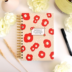 May include: A spiral-bound weekly planner with a beige cover featuring a red floral pattern and white polka dots. The center text reads "WEEKLY PLANNER" with Japanese characters below. The planner has gold-colored metal wire binding.