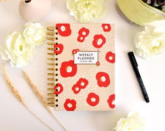 May include: A spiral-bound weekly planner with a beige cover featuring a red floral pattern and white polka dots. The center text reads "WEEKLY PLANNER" with Japanese characters below. The planner has gold-colored metal wire binding.