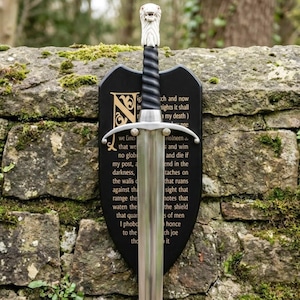 Puede incluir: Una espada de metal con hoja plateada y empuñadura negra de textura en espiral, rematada con un pomo en forma de cabeza de lobo blanca. La espada está montada sobre una placa negra en forma de escudo con texto dorado que incluye la palabra Night.