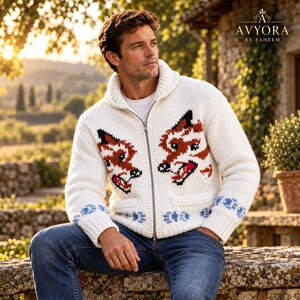 May include: A white knit zip-up cardigan featuring a shawl collar and two fox head graphics in orange, black, and white. The hem and cuffs are decorated with a blue floral pattern. The text AVYORA BY FAHEEM is visible in the top right corner.