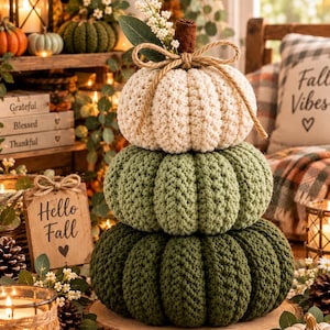 May include: A stacked set of three crocheted pumpkins in cream, sage green, and dark forest green. The top pumpkin features a brown stem, a jute twine bow, and a small sprig of white flowers. The pumpkins are textured with vertical ridges.