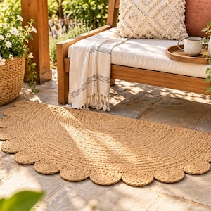 May include: A semi-circular rug crafted from natural tan woven jute fibers, featuring a decorative scalloped edge design. The rug has a flat, braided texture and is positioned on a stone patio surface, showcasing a consistent light brown color.