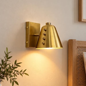 May include: A wall-mounted lamp featuring a brushed brass finish with a conical shade. The fixture includes a rectangular backplate and a series of four small, spherical decorative studs arranged vertically along the side of the metal cone.