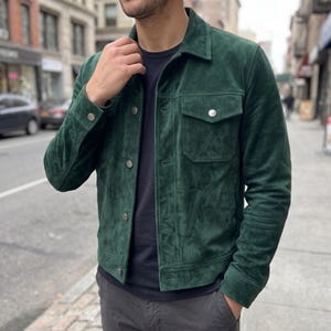 May include: A dark green suede jacket featuring a classic collar, long sleeves with buttoned cuffs, and a front button closure. The garment includes two chest pockets with buttoned flaps and a smooth, matte texture throughout the material.